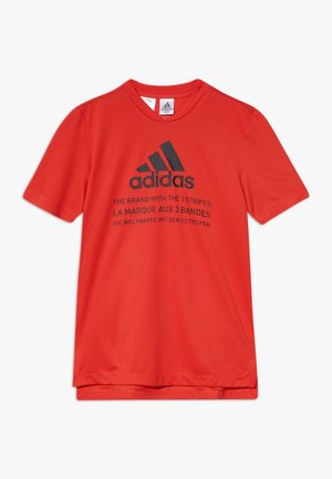 T-shirt de sport rouge à manches courtes Adidas avec logo noir et texte multilingue promouvant les trois bandes de la marque à l'avant.