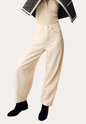 Person trägt hoch taillierte, weit geschnittene cremefarbene Hose, beigefarbenes Henley-Shirt, schwarze Stiefeletten und hält eine gemusterte Jacke über den Schultern.