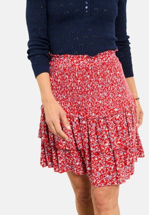 Woman wearing a navy blue long-sleeve top and a red floral tiered skirt, with hands resting near the skirt's pleats.