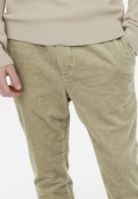 Pantalones de pana verde oliva con costillas verticales, que presentan un corte relajado y bolsillos laterales, combinados con un suéter de canalé beige claro.