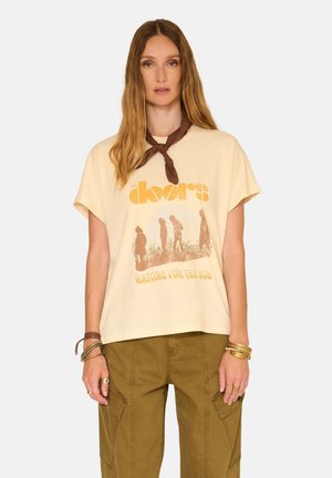 Femme portant un t-shirt graphique The Doors jaune clair, un pantalon cargo marron, un foulard autour du cou et des bracelets, debout devant un fond uni.