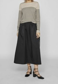 Model nosząca beżową i czarną paskowaną bluzkę z długim rękawem, zestawioną z czarną, długą spódnicą o fakturze i czarnymi sandałami na obcasie.