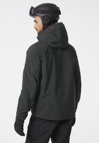 Zwarte waterdichte skijas met een capuchon, elastische manchetten en een matte afwerking. Draagt een zwarte helm en handschoenen, en toont een praktische vormgeving.