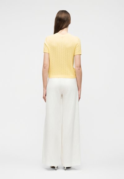 Γυναίκα που στέκεται με την πλάτη προς τα εμπρός, φοράει κοντομάνικη πλεκτή κίτρινη μπλούζα και άσπρο παντελόνι με φαρδιά πόδια, με τακούνια, πάνω σε απλό λευκό φόντο.