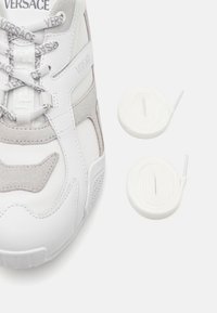 Zapatilla blanca con detalles en ante gris, que presenta un diseño texturizado, un logo prominente y cordones blancos. Se incluyen dos cordones redondos blancos adicionales.