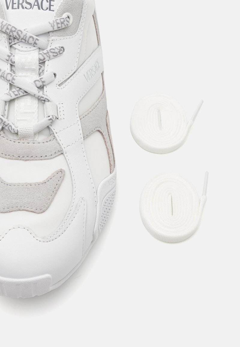 White sneaker with gray suede accents, featuring a textured design, prominent logo, and white laces. Two additional white round laces included.