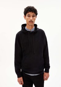Sweat-shirt à capuche noire en tissu doux, doté d'une poche avant, de cordons de serrage et d'un petit logo sur la poitrine. Superposé sur une chemise à col.