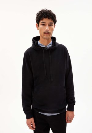 Black hooded sweatshirt made of a soft fabric, featuring a front pocket, drawstrings, and a small logo on the chest. Layered over a collared shirt.