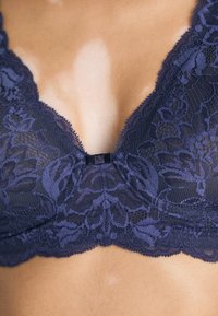 Close-up van een persoon die een donkerblauwe kanten beha draagt met bloemmotieven en een klein decoratief strikje in het midden.