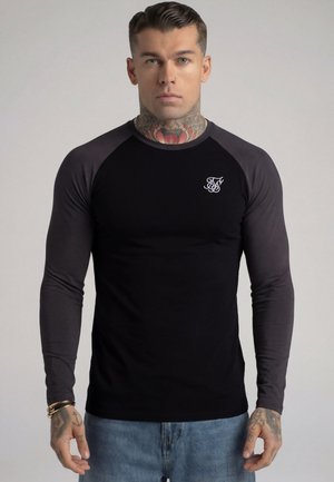 Mann med tatoveringer på nakken og hendene, iført en svart langermet skjorte med hvit logo og blå jeans, vendt mot kamera foran ensfarget bakgrunn.