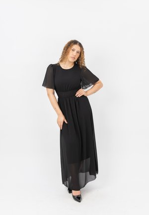 Femme aux cheveux bouclés portant une robe maxi noire transparente et des talons noirs, debout avec une main sur la hanche sur un fond blanc.