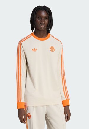 Man met dreadlocks, gekleed in een beige trainingspak met feloranje strepen, boorden en logo's op de borst en broek, staand tegen een effen achtergrond.