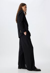 Blazer noir ajusté avec des boutons dorés, associé à un pantalon noir ample. Le modèle se tient de profil, mettant en valeur le design épuré et structuré ainsi que l'ajustement.