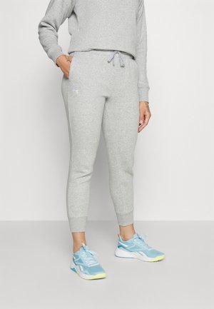 Personne portant un pantalon de survêtement gris clair et un sweat assorti, avec des chaussures de sport bleu clair, debout avec une main dans la poche.