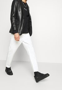 Černé kožené sako zkombinované s černým tričkem a bílými kalhotami. Černé tenisky doplňují vzhled. Modelka kráčí.