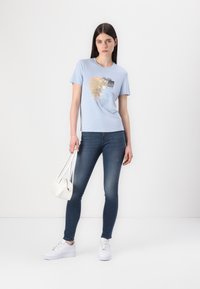 Lichtblauwe T-shirt met een grafische print, gecombineerd met donkere jeans en witte sneakers. Het model houdt een kleine witte handtas vast.