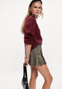 Burgundský pletený sveter s okrúhlym výstrihom, skombinovaný so záhybovou béžovou kockovanou sukňou. Model drží čiernu kabelku s ramenným popruhom.