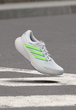 Chaussure de course Adidas Supernova blanche avec des bandes vert néon, capturée en plein saut au-dessus d'une route goudronnée avec des marquages blancs.