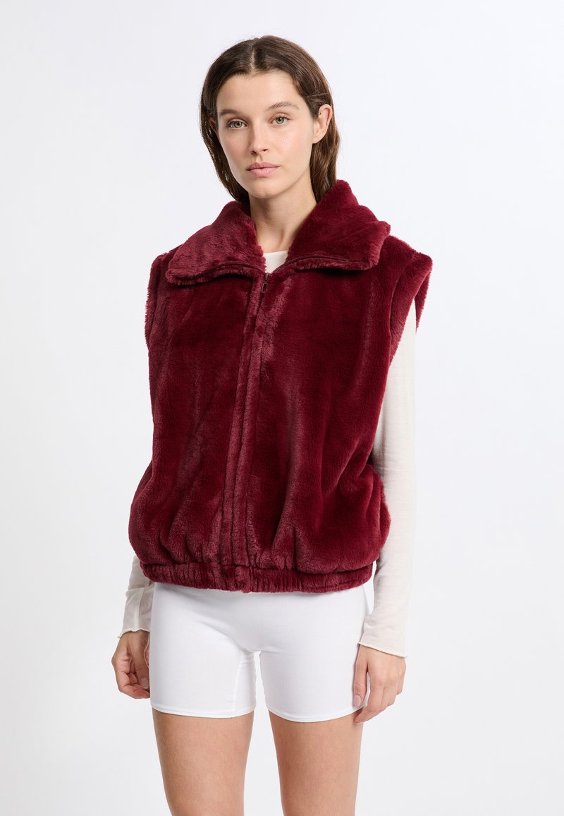 Gilet in finta pelliccia bordeaux con alto colletto, chiusura con zip e orlo elastico; indossato sopra una maglietta a maniche lunghe chiara e pantaloni corti bianchi.