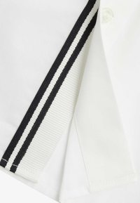 Camicia bianca con un bordo a righe nere lungo il colletto, dotata di dettagli con bottoni e una consistenza del tessuto liscia.