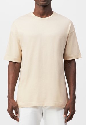 Mann trägt ein schlichtes beige T-Shirt mit kurzen Ärmeln und weiße Hose, steht vor einem neutralen grauen Hintergrund.