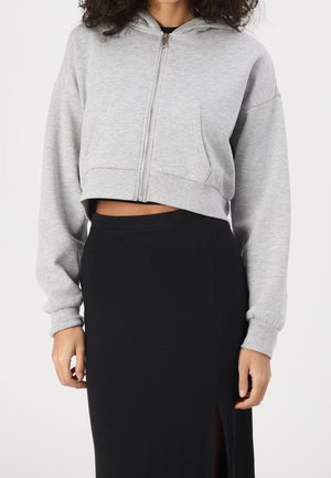 Vrouw die een lichtgrijze cropped zip-up hoodie met voorzakken draagt en een zwarte hoge taille rok met een voorwaartse split.