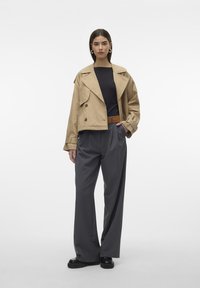 Beige lühendatud trench coat suure kraega, kahe ridadega disain, koos hallide volditud laia säärega pükste ja mustade kingadega.