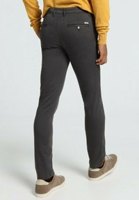 Pantalon slim gris foncé avec une texture lisse, doté de deux poches arrière et d'un détail avec un bouton. Porté avec des baskets marron clair.