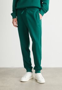 Grønne joggebukser med snor i livet, sidelommer og subtile logo detaljer. Kombinert med hvite sneakers på en nøytral bakgrunn.