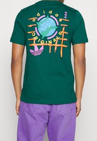 Grön bomullströja med en färgglad globdesign, med orange linjer och text som lyder "adidas Originals", samt en lila logotypdetalj.