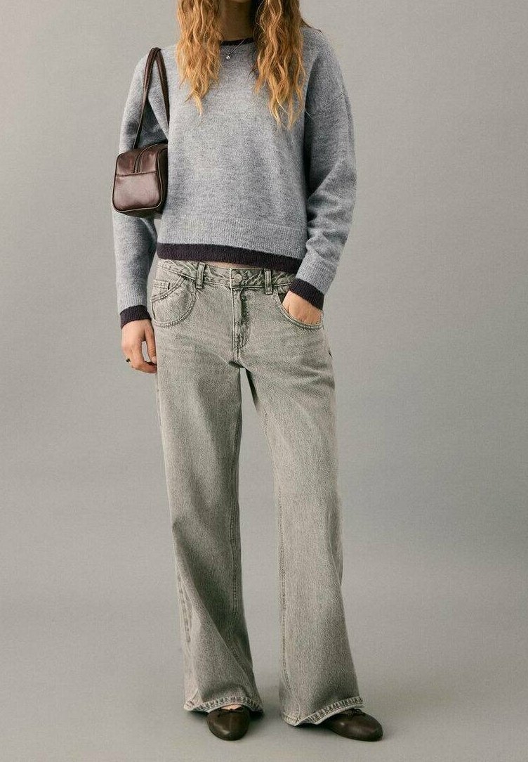 Jeune femme portant un pull gris, un jean ample gris clair, des chaussures marron foncé et portant un petit sac à épaule marron foncé.