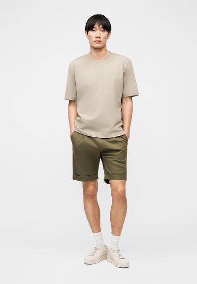 Νέος άνδρας που στέκεται με τα χέρια στις τσέπες, φοράει μπεζ μπλουζάκι, χακί σορτς, λευκές κάλτσες και μπεζ αθλητικά παπούτσια σε λευκό φόντο.