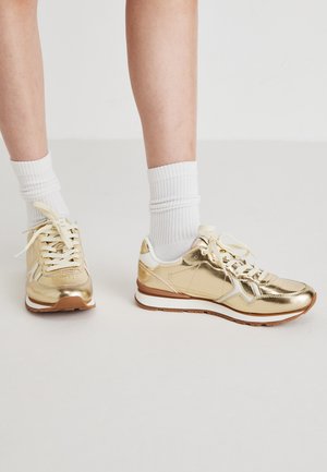 Zelta metāla teniski ar baltajām akcentiem, ar plakanu auklu, mīkstām zolēm un struktūrētām paneļiem, kombinēti ar baltām ribotām zeķēm.