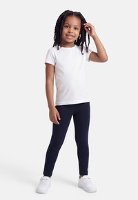 Χαμογελαστή κοπέλα με πλεξούδες, ντυμένη με λευκό T-shirt, μπλε σκούρο κολάν και λευκά αθλητικά παπούτσια, στέκεται αγγίζοντας τα μαλλιά της μπροστά σε απλό φόντο.