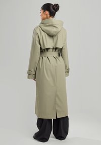 Pitkä oliivinvihreä sadetakki, jossa on huppu, vyötäröllä vyö ja poimutettu takakappale. Siinä on pitkät hihat ja halkiohelma. Tasainen, vedenpitävä materiaali.