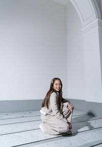 Jasny beżowy garnitur z krótka marynarką i szerokimi spodniami, w połączeniu z białą koszulą. Siedzi na marmurowej posadzce z szarymi akcentami.