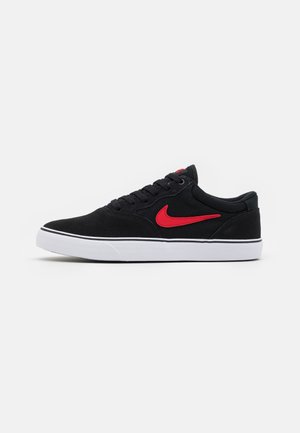 Lavtops sort sneaker med rød Nike-swoosh og hvid sål, set fra siden på en ensfarvet lys grå baggrund.