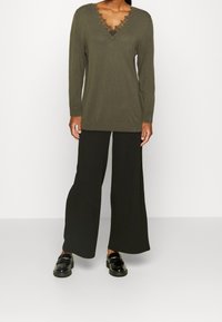 Oliivinvihreä, pitkähihainen paita pitsireunaisella v-pääntiellä, yhdistetty leveälahkeisiin mustiin housuihin ja mustiin loafer-kenkiin. Yksinkertainen, teksturoitu muotoilu.