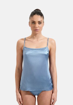 Woman with dark hair in a bun wearing a shiny light blue satin camisole top and matching underwear, standing against a plain light background.