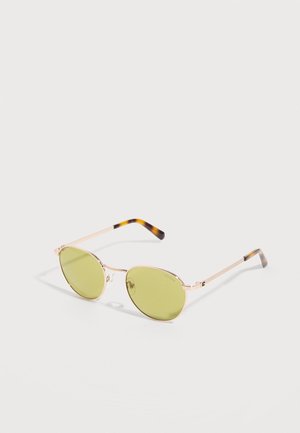 Lunettes de soleil rondes avec monture en métal doré, verres teintés jaunes et branches à motif écaille sur fond blanc.