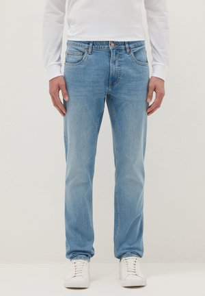 Hombre con jeans de pierna recta azul claro, camisa blanca de manga larga y zapatillas blancas, posando frente a un fondo liso.