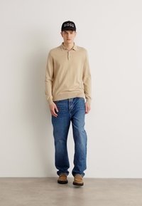 Polo a maniche lunghe beige, jeans in denim blu, scarpe marroni e un cappellino nero con lettering bianco "BOSS". Outfit semplice e casual.
