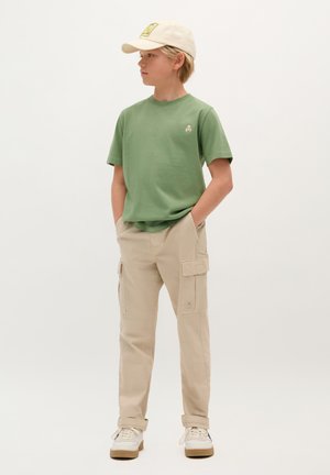 T-shirt verde in cotone con un piccolo logo, pantaloni cargo beige con tasche laterali e un cappellino color crema con logo. Sneakers bianche con suole beige.