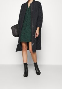 Manteau long matelassé noir avec des boutons-pression, superposé à une robe verte à motifs avec des boutons. Des bottines noires à la cheville complètent le look.