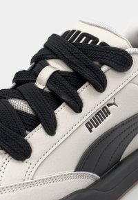 Sapatilhas de couro branco com atacadores e detalhes pretos. Apresentam um design texturizado, um proeminente logótipo da Puma e uma faixa lateral cinza.