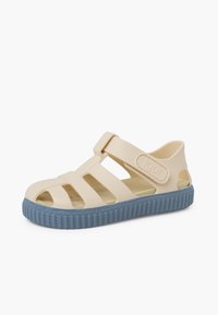Beige rubber sandal with open sides and adjustable strap, featuring a textured blue sole and embossed logo on the strap.