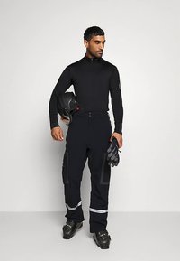 Vīrietis melnā slēpošanas tērpā, kurš tur helmīti un cimdus, valkā slēpošanas zābakus un skatās uz sāniem pret vienkāršu fona.