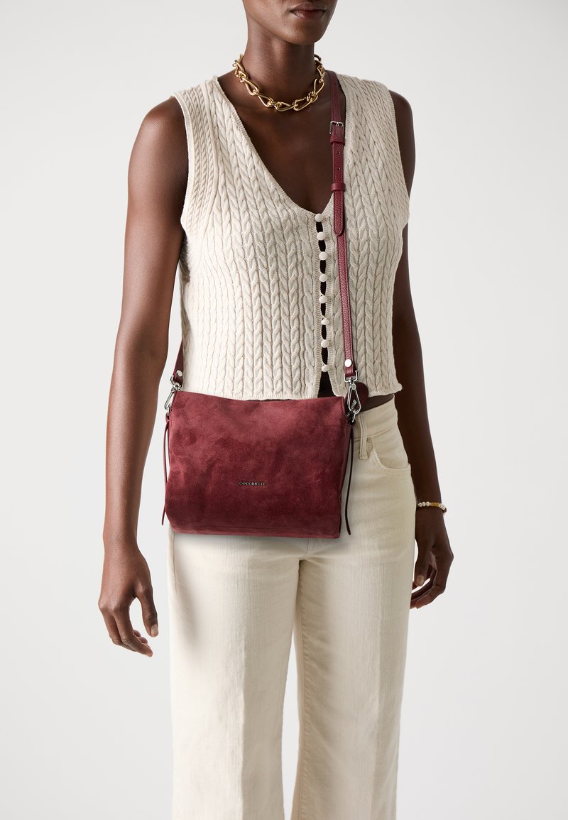 Burgundy suede crossbody bag with adjustable strap, featuring a smooth, matte texture and subtle logo detail on the front.