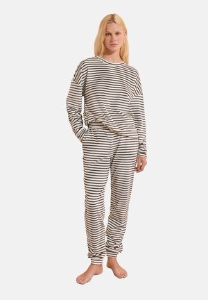 Femme debout, pieds nus, portant un haut à manches longues et un pantalon assortis avec des rayures horizontales noires et blanches sur un fond uni.