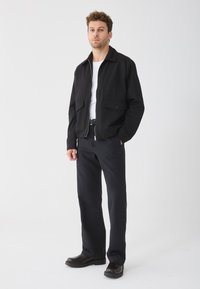 Juodas užtrauktuku užsegamas švarkas su apykakle, dviem priekinėmis kišenėmis, derinamas su juodomis kelnėmis ir tamsiais batais. Audinys atrodo lygus ir lengvas.